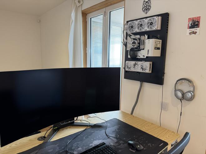 Photograph of the final product. There&rsquo;s a corner of the desk showing part of a keyboard, mouse and monitor and hanging from the wall: the computer. it has a black support and white components. Only motherboard, graphics card PSU and CPU radiator are visible
